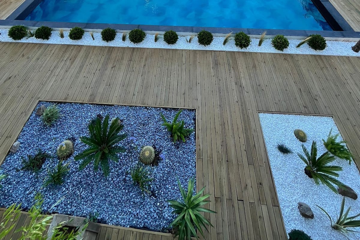 Een rechthoekig zwembad met een houten terras eromheen, met ligbedden en een zitgedeelte onder een rieten dak.