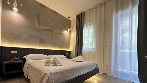 A modern hotel room with a large bed, a bedside table, a wall-mounted light fixture, and a window with curtains. - Marilu Vacanze (Bari)