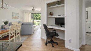 A modern living room with a dining area, a grey sofa, a black office chair, and a TV on a white shelf. - Luxury 2BR Shipyard Villa: Golf Views, Heated Spa & Walk to Beach (Hilton Head Island)