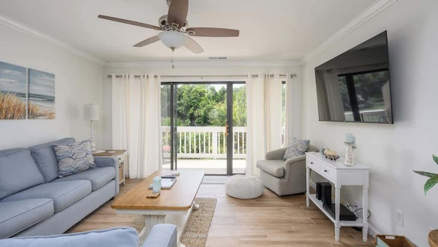 A modern living room with a ceiling fan, a large window with curtains, a flat-screen TV, a sofa, a coffee table, and a side table with decorative items. - Luxury 2BR Shipyard Villa: Golf Views, Heated Spa & Walk to Beach (Hilton Head Island)