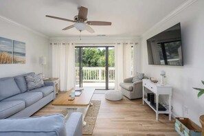 Una sala de estar moderna con ventilador de techo, un ventanal con cortinas, televisor de pantalla plana, sofá, mesa de centro y una mesita auxiliar con objetos decorativos.