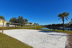 A sandy beach volleyball court with a net, surrounded by a lake, palm trees, and a building in the background.