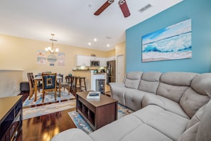 A modern living room with a beige sofa, wooden dining table, and chairs. There is a kitchen area with white cabinets and a microwave. A painting of a beach scene is on the wall.