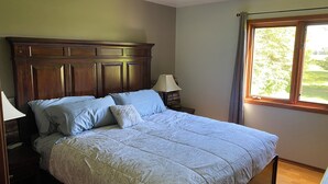 Un dormitorio con cabecera de madera, cama con ropa de cama floral, lámpara de noche y ventana con vista a la vegetación.