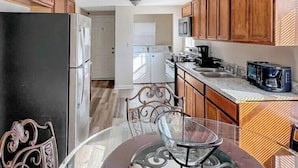 A compact kitchen with wooden cabinets, a refrigerator, a microwave, a coffee maker, and a dining area with a glass table and metal chairs. - Great Vacation Escape (Savannah)
