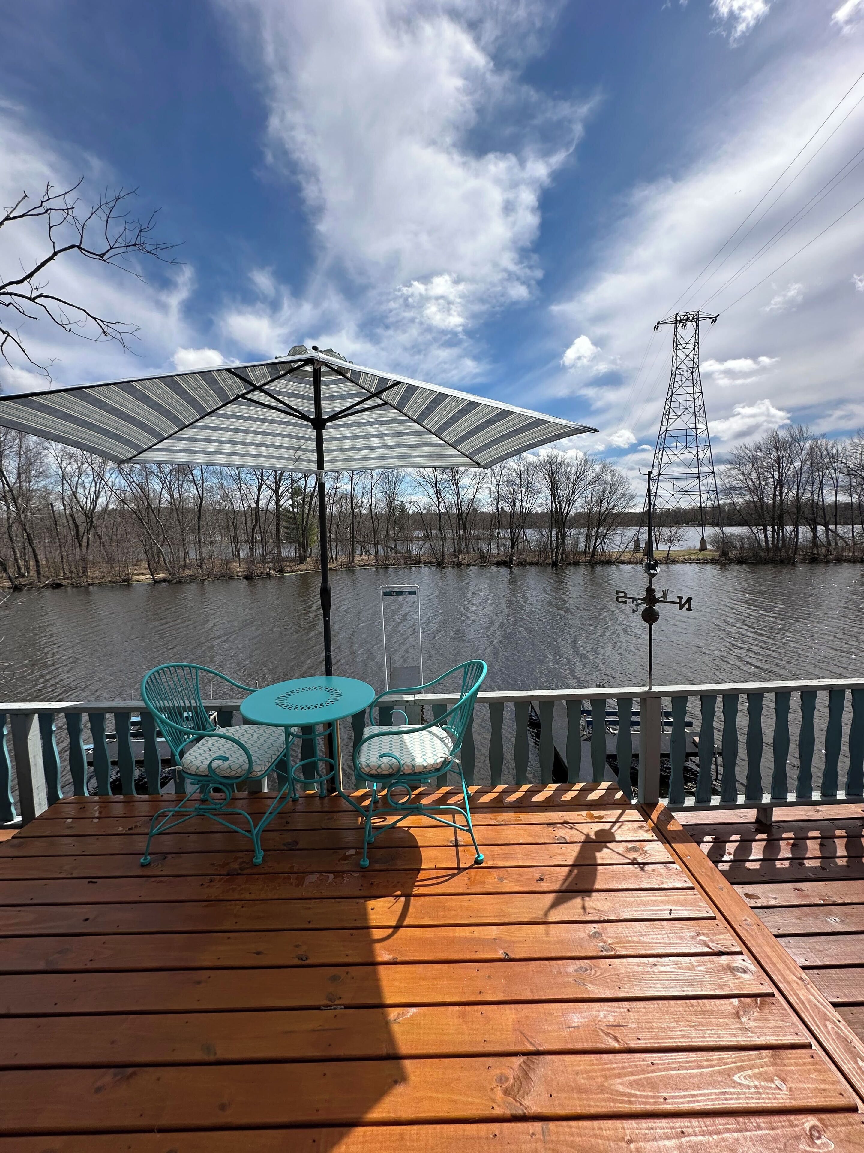 Una terraza de madera con dos sillas turquesa y una mesa, con vista a un cuerpo de agua y una torre de líneas eléctricas al fondo.