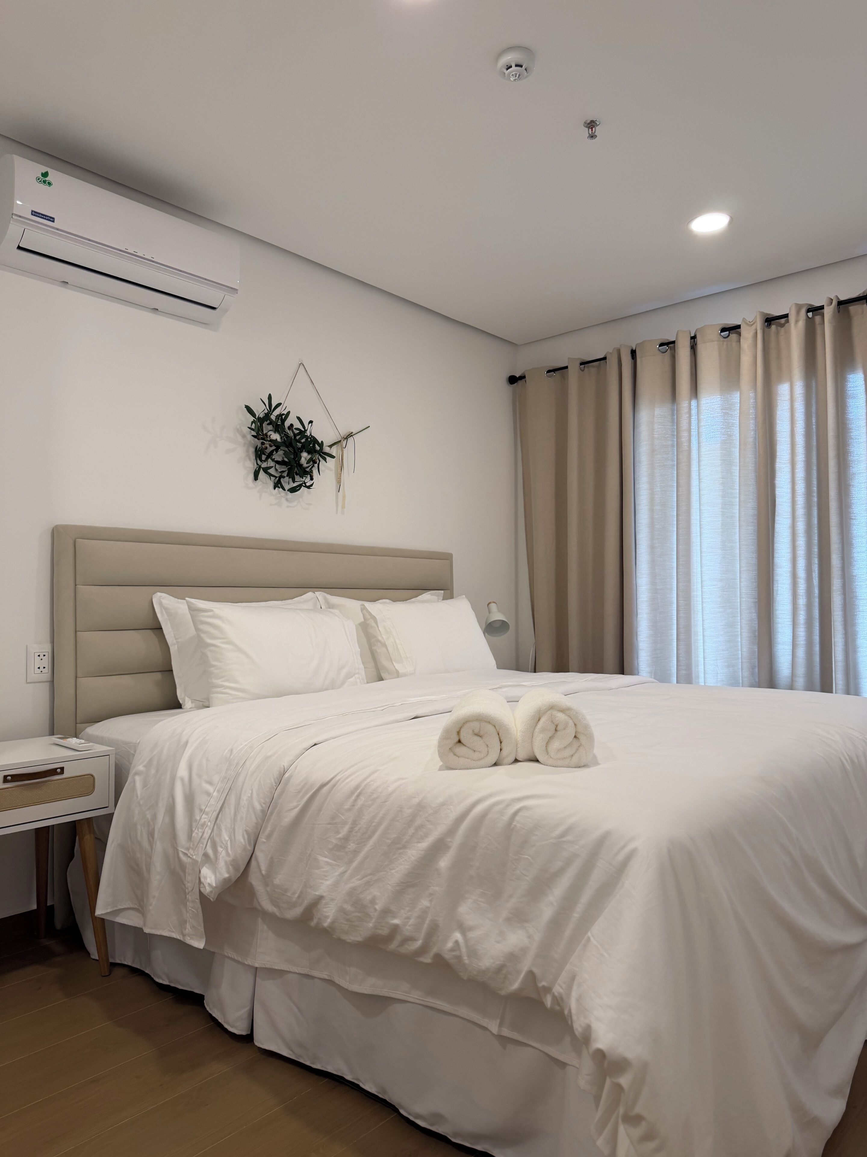 A neatly made bed with white linens and rolled towels, a bedside table with a lamp, and an air conditioning unit on the wall.