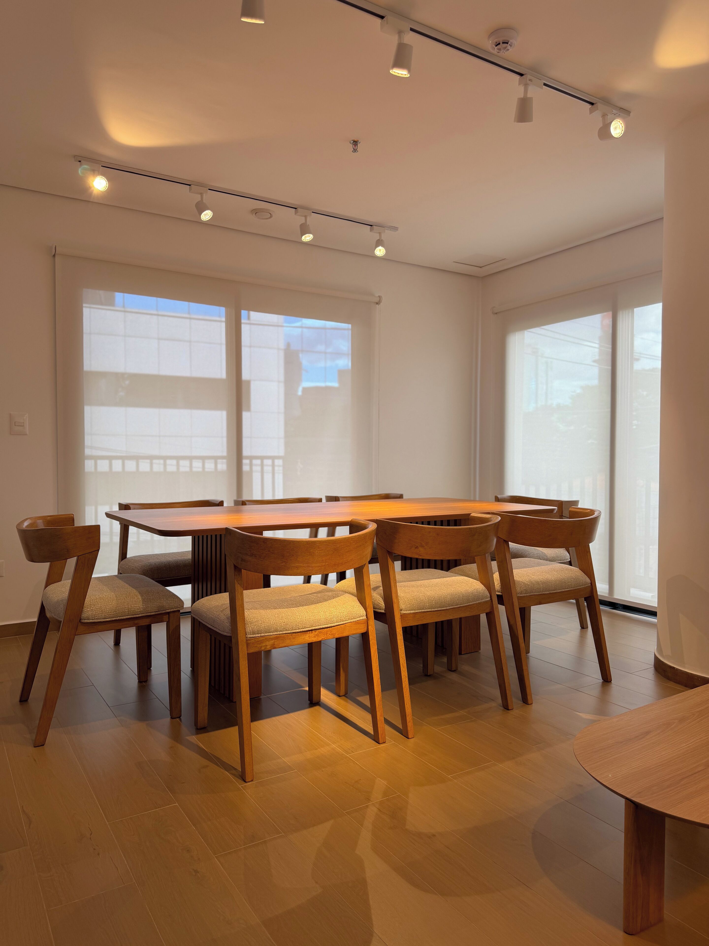 Uma sala de jantar com mesa e cadeiras de madeira, janelas amplas e luminárias no teto.