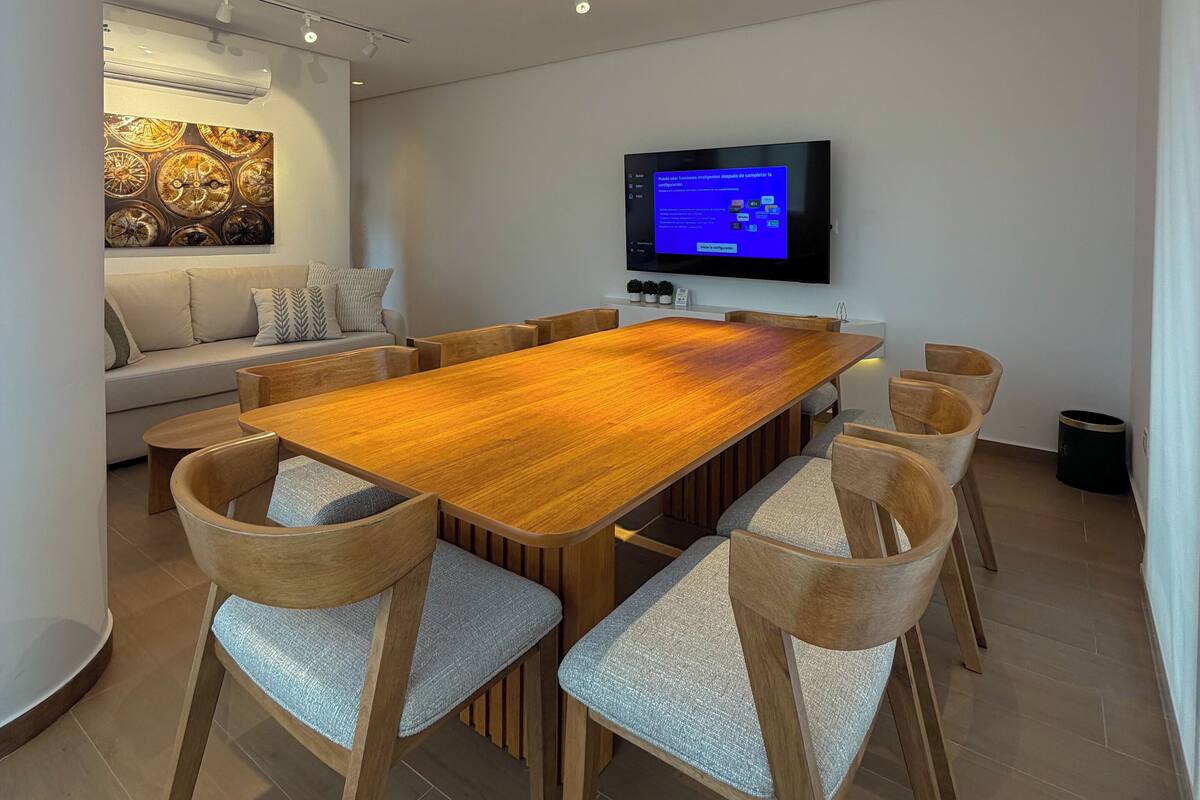 Un comedor moderno con una larga mesa de madera, seis sillas tapizadas, un televisor de pantalla plana montado en la pared e iluminación empotrada en el techo.