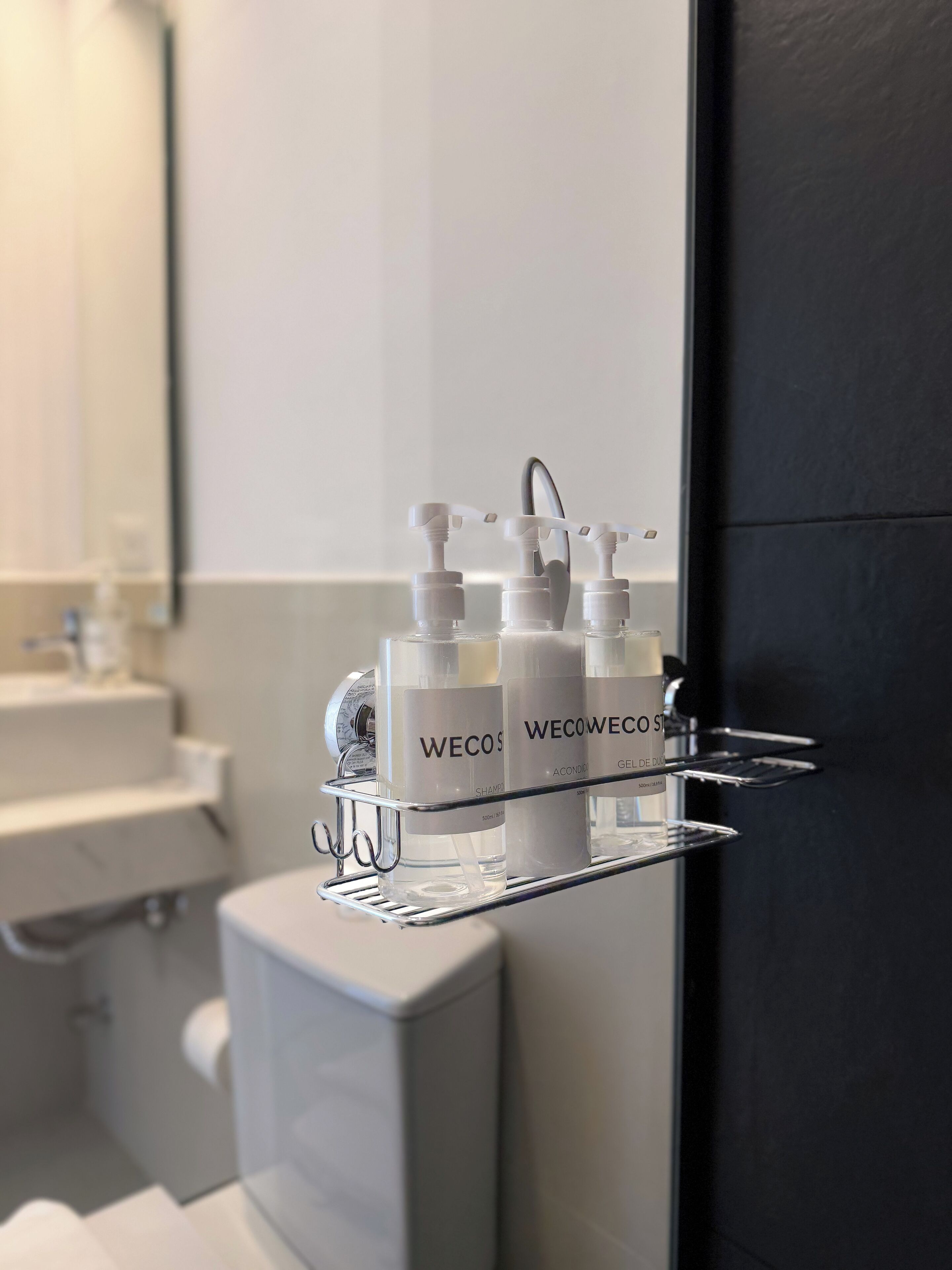Une salle de bain avec un lavabo blanc, un miroir et une étagère sur laquelle sont posées trois bouteilles de shampoing WeCos.