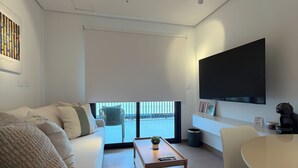A modern living room with a sofa, coffee table, and a dining area with chairs. There is a large window providing a view outside.