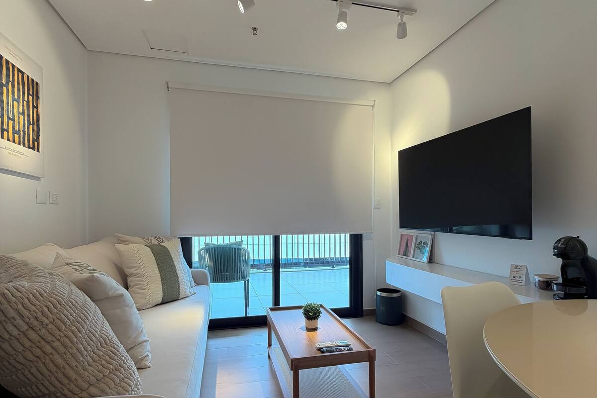Una sala moderna con sofá, mesa de centro y zona de comedor con sillas. Una ventana grande ofrece vistas al exterior.