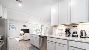 Moderni keittiö, jossa on valkoiset kaapit, astianpesukone, kahvinkeitin ja tehosekoitin.