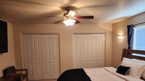 A bedroom with a bed, a desk, two doors, a ceiling fan, and a window with curtains.