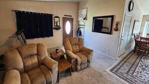 A living room with two brown armchairs, a small wooden table, a treadmill, a door with a mirror, and a patterned rug.