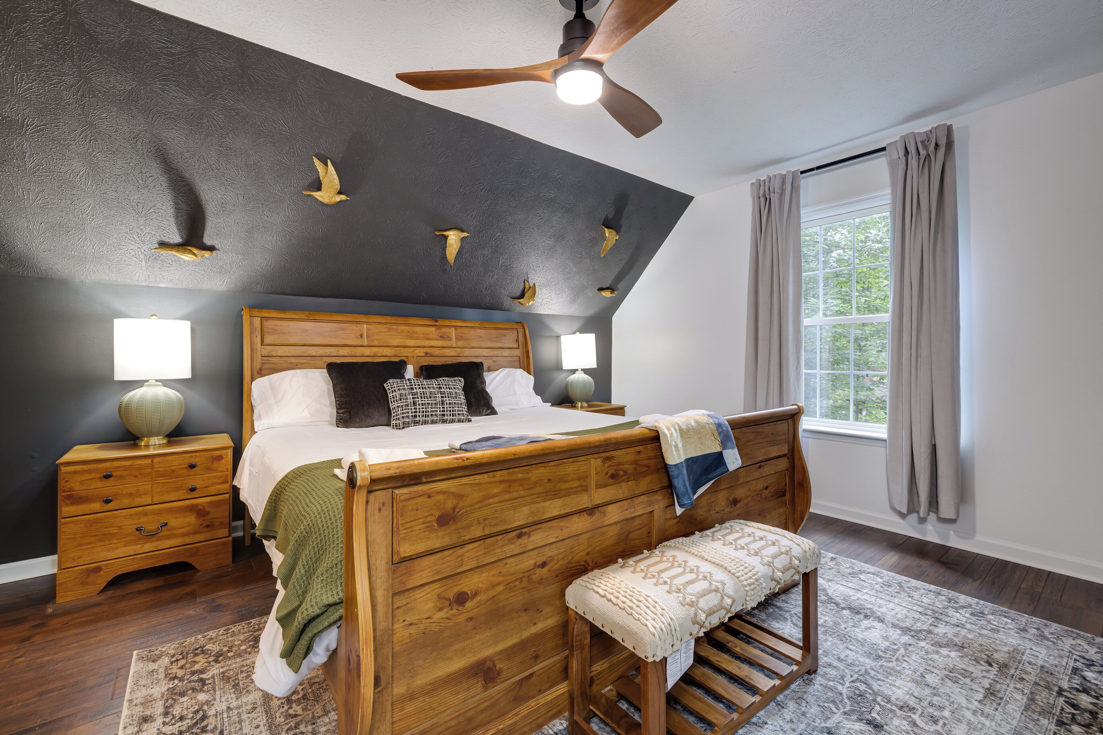 A bedroom with a wooden bed, nightstands, a bench, a ceiling fan, and a window with curtains.