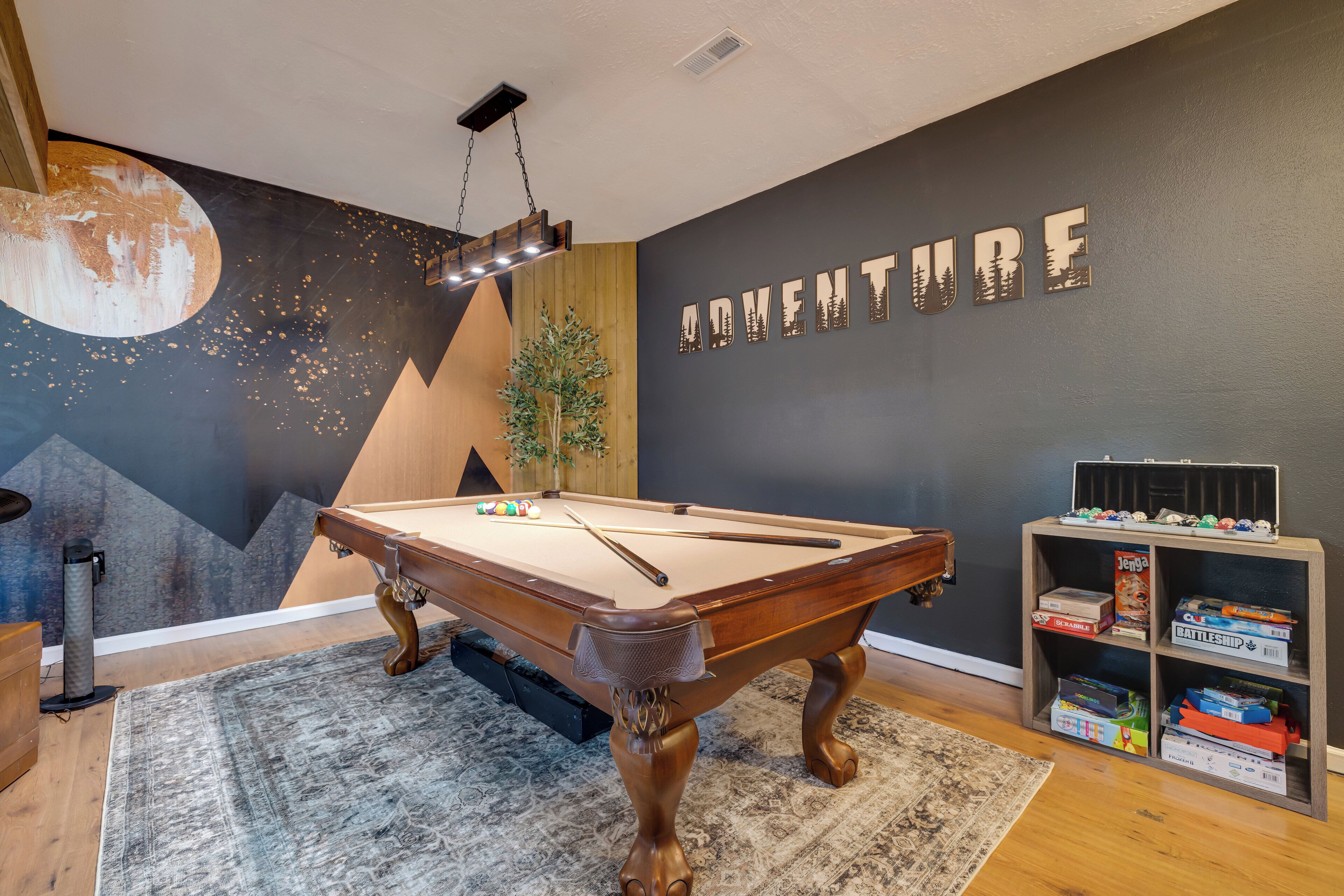 A pool table in a room with a 'ADVENTURE' wall mural, a bookshelf with board games, and a large rug.