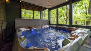 A hot tub with a wooden deck and a view of a forest through large windows.