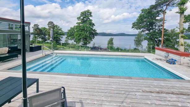 Ein Schwimmbad mit Holzterrasse, Liegebereich und Blick auf einen See und Berge.