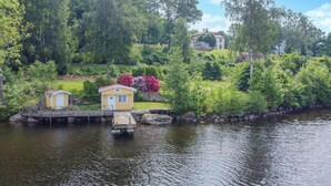 Ein Haus am Seeufer mit Steg, einer kleinen gelben Lagerhütte und einem großen Haus im Hintergrund.