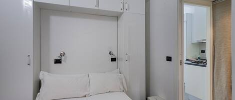 A bedroom with a bed, white curtains, a small stool, and a view of a kitchen through an open door.