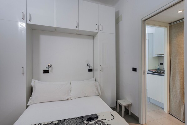 Une chambre à coucher avec un lit, des rideaux blancs, un petit tabouret et une vue sur une cuisine par une porte ouverte.