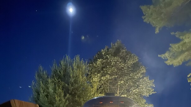 A camper van parked at night with a campfire in front, surrounded by wooden furniture and trees.