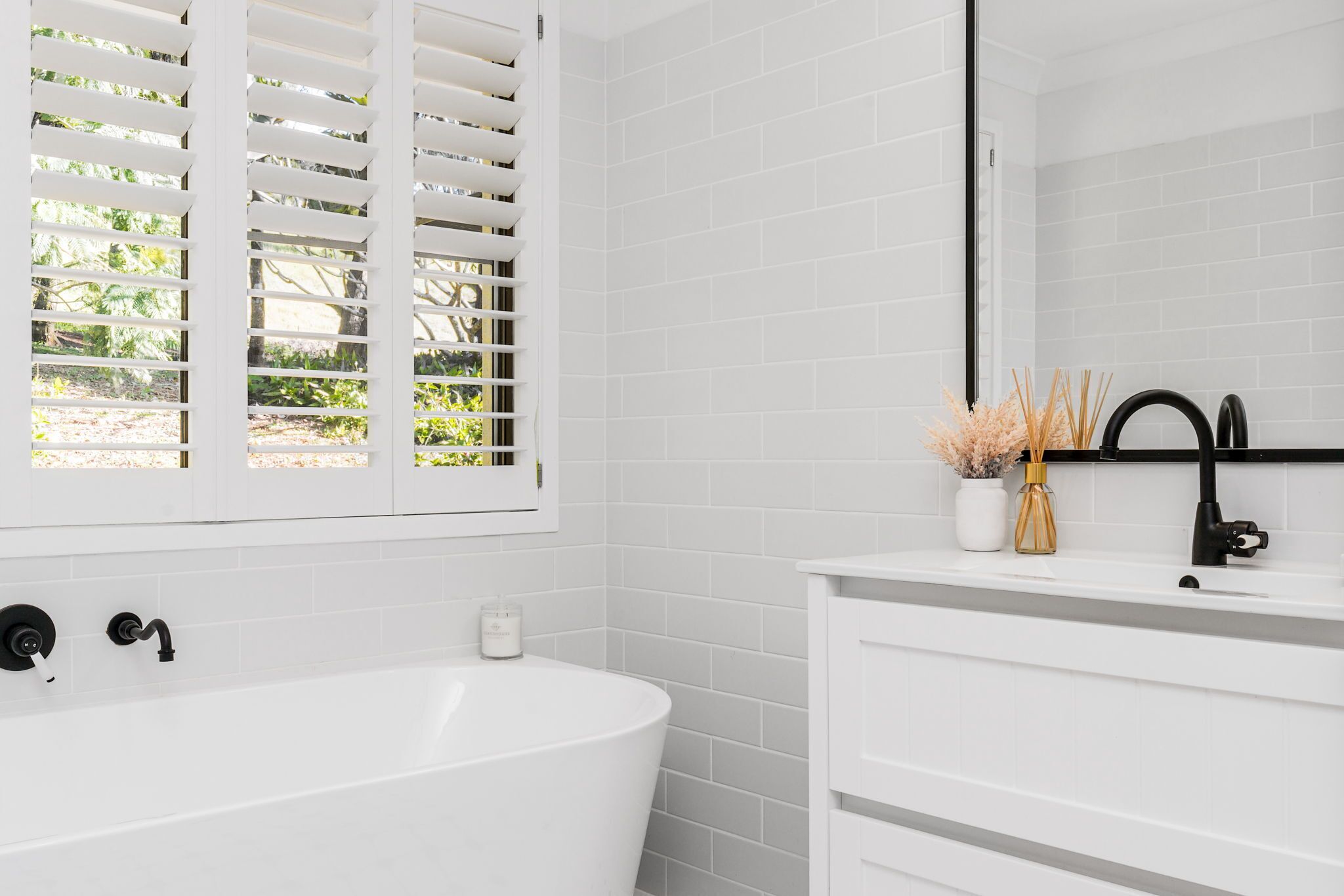 A modern bathroom with a freestanding bathtub, white tiled walls, a large mirror, and a window with white blinds.