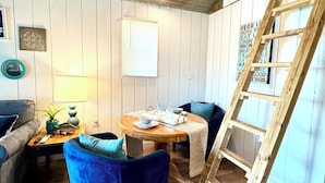A rustic room with wooden floors, a round wooden table, two blue velvet armchairs, a wooden ladder, and a white wall with a window.