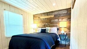 A bedroom with a wooden ceiling, a bed with a blue bedspread, two bedside lamps, and a window with blinds.