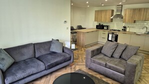 A modern living room with two grey sofas and a black coffee table, a kitchen with wooden cabinets and stainless steel appliances, and a herringbone-patterned wooden floor. - Spacious City Living / Free Parking (Birmingham)