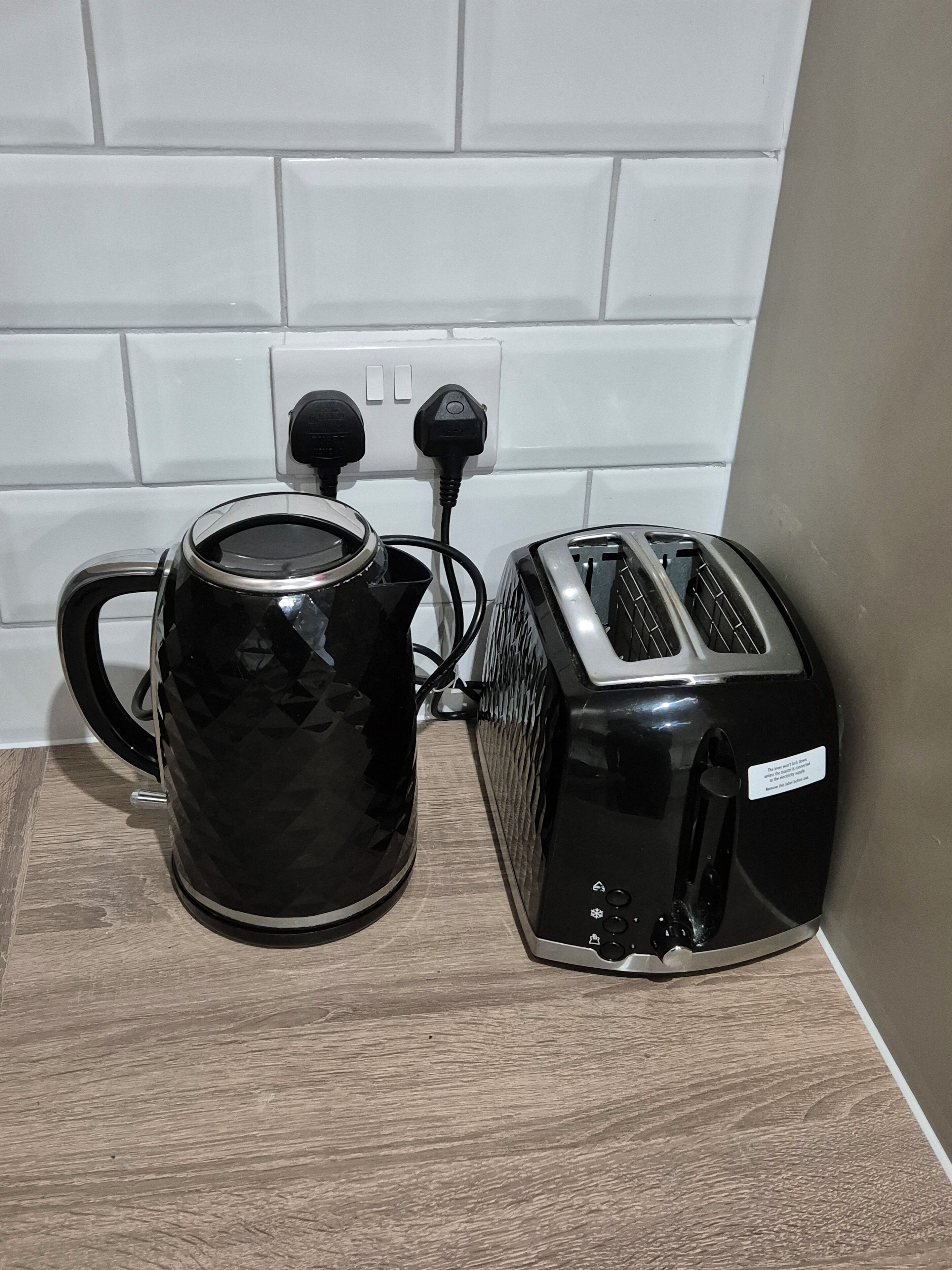 Eine Kaffeemaschine und ein Toaster in Schwarz stehen auf einer Holzauskładfläche, verbunden mit einer weißen Steckdose.