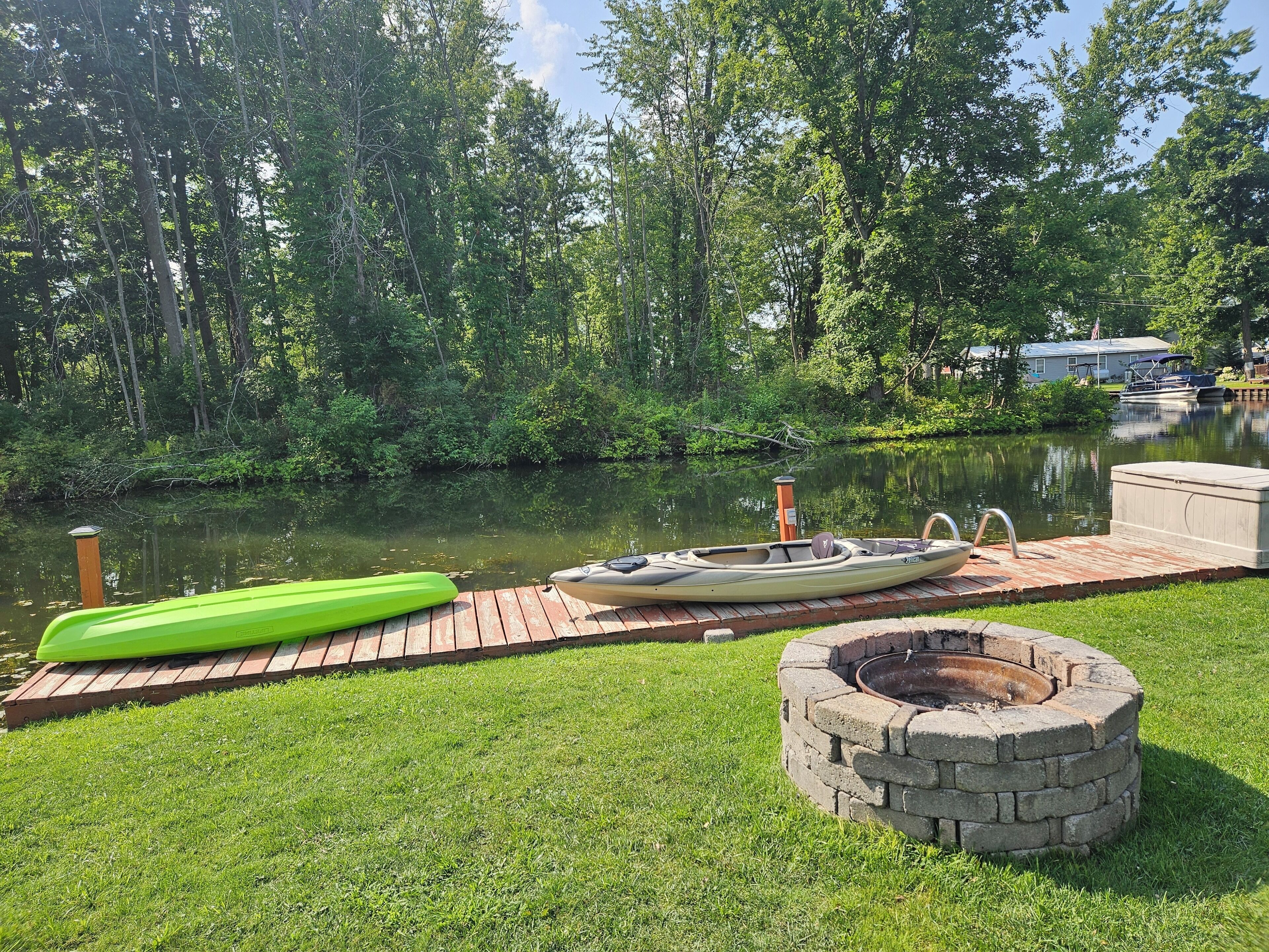 A wooden dock with a kayak and a green paddleboard, a stone fire pit, and a calm river surrounded by trees.