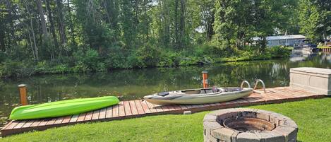 A wooden dock with a kayak and a green paddleboard, a stone fire pit, and a calm river surrounded by trees.