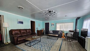 Una sala de estar con un sofá de cuero marrón, una mesa de centro, un tapete estampado, piso de madera, un ventilador de techo, una chimenea y un televisor.