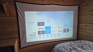 Proyección de una interfaz de dispositivo en una pared, mostrando una fecha, hora y una lista de aplicaciones, incluyendo YouTube y Netflix.