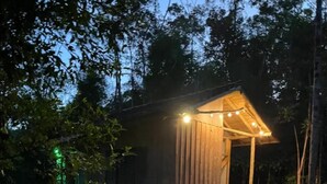 Una pequeña cabaña de madera con un porche, rodeada de árboles y césped.