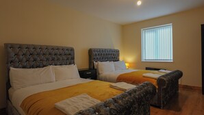 A hotel room with two beds, each with a tufted headboard and white bedding. There is a window with blinds, a bedside table with a lamp, and a chair with a cushion. - The Blissful Heaven (Manchester)