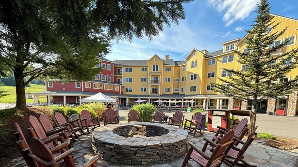 Un hogar de piedra rodeado de sillas Adirondack, con un edificio al fondo que presenta paredes exteriores amarillas y rojas.