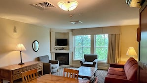 A hotel room with a wooden dining table, two armchairs, a red sofa, a TV mounted above a fireplace, and a window with curtains.