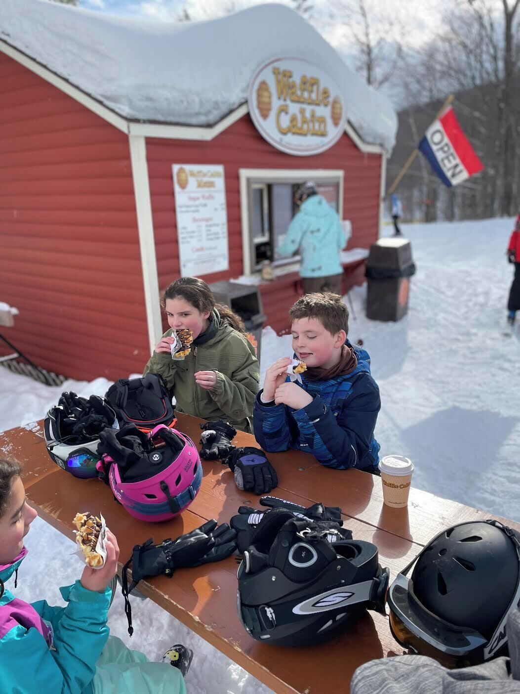 Un grupo de personas comiendo en una mesa al aire libre, con nieve en el suelo, un edificio rojo con un letrero que dice “Waffle Cabin” al fondo, y una bandera que indica que el establecimiento está abierto.