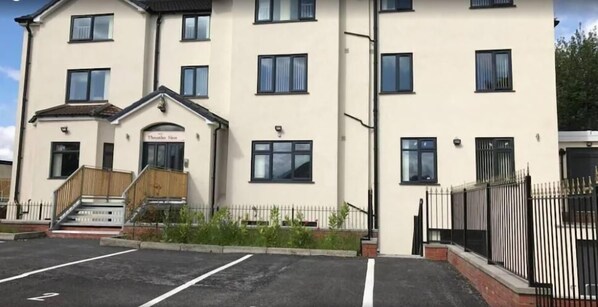 Un edificio a più piani con facciata piatta, un ingresso principale con gradini e un'area di parcheggio di fronte.