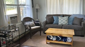 A living room with a sofa, a coffee table, a wicker chair, a floor lamp, and a window with blinds. - Family Friendly Lake View Home. Only steps away from Houghton Lake. (Houghton Lake)