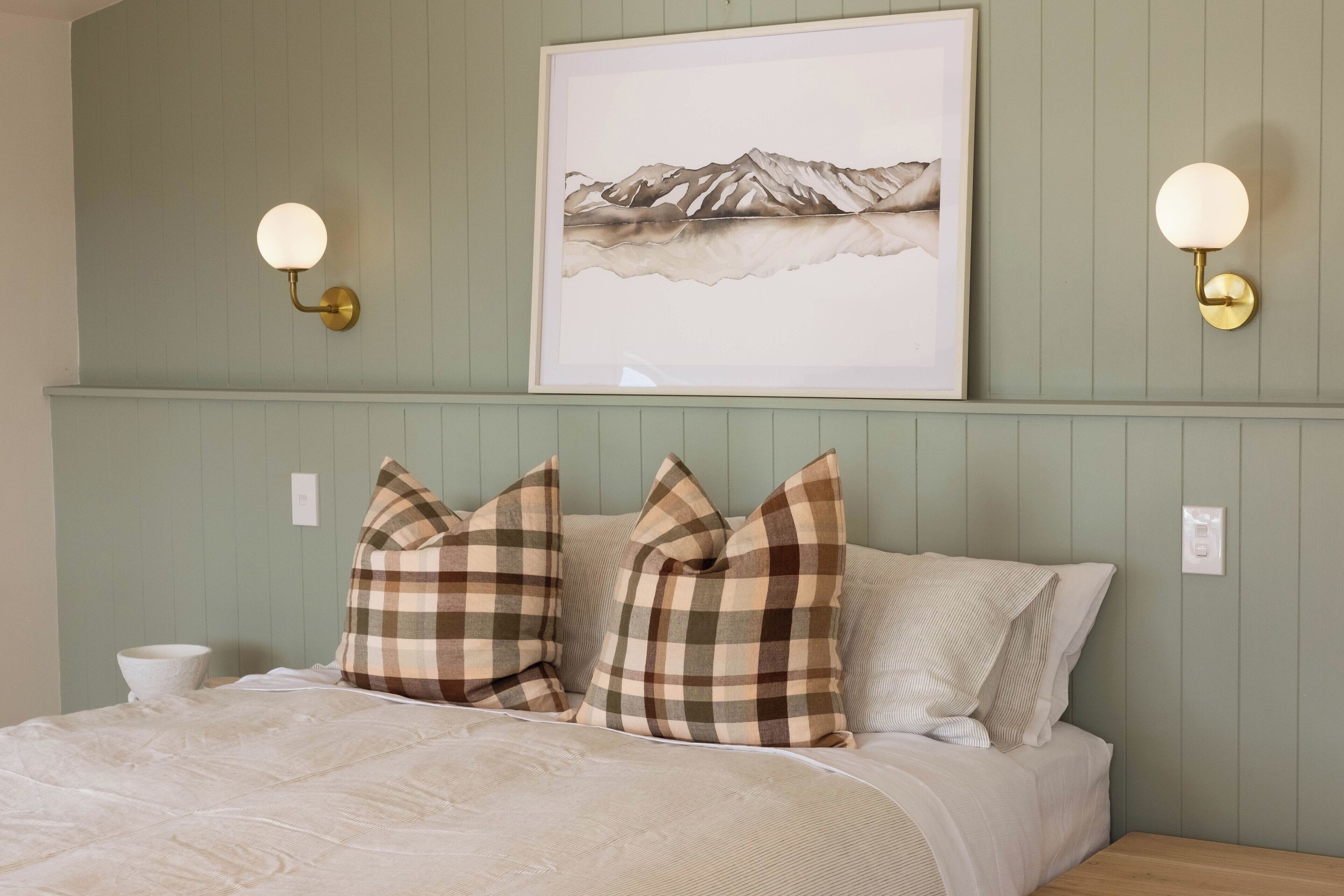 Ein Schlafzimmer mit einem Bett, karierten Kissen, einem gerahmten Bild an der Wand und Wandlampen.