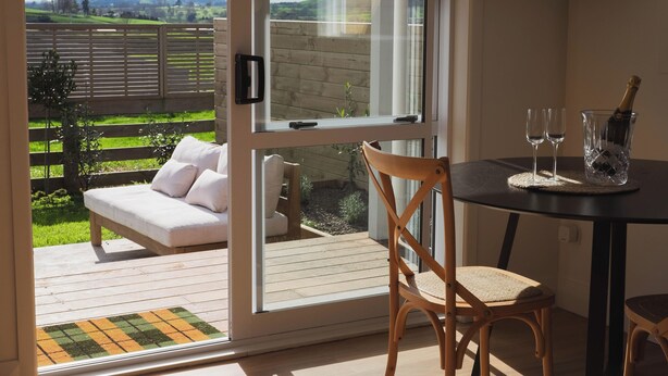 A room with a sliding glass door leading to an outdoor deck, a wooden chair, a small table with two glasses and a bottle of champagne, and a TV mounted on the wall. - Willow View Cottage with beautiful country views - Near Waitomo (Otorohanga)
