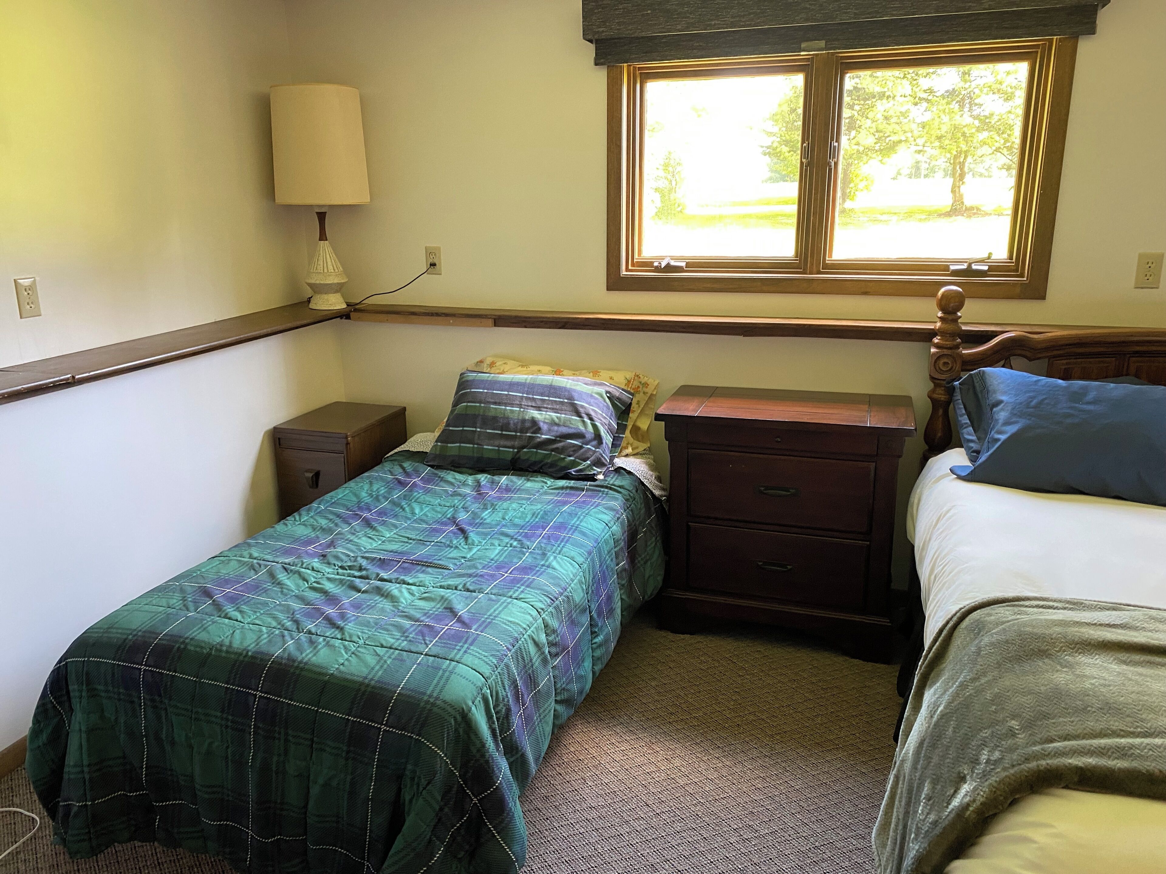 Une chambre avec deux lits, une table de chevet, une commode, une lampe et une fenêtre donnant sur des arbres.