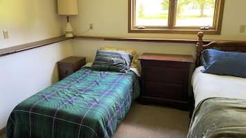 Une chambre avec deux lits, une table de chevet, une commode, une lampe et une fenêtre donnant sur des arbres.