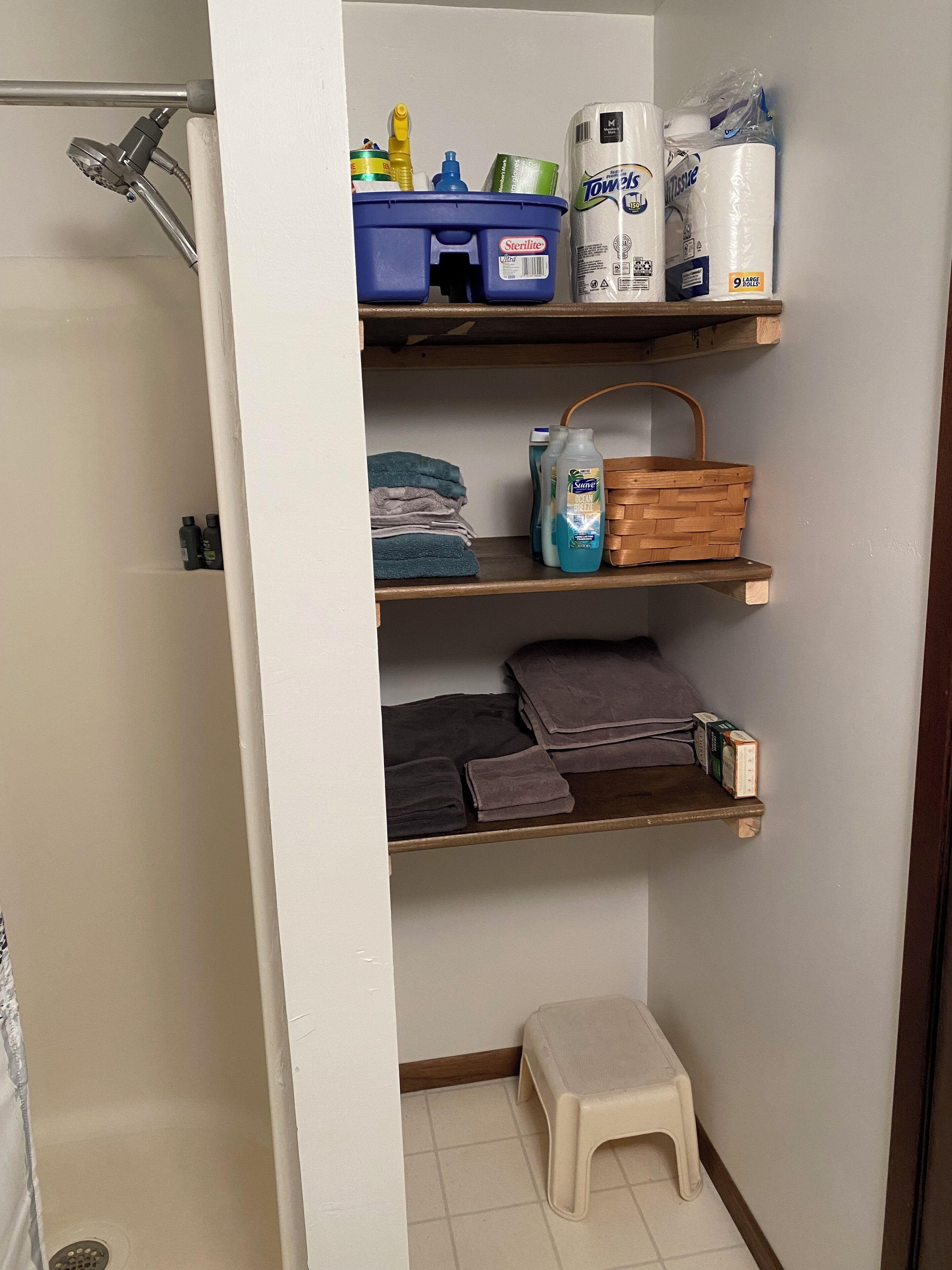 Een badkamer met een douche, planken met toiletartikelen, handdoeken en een klein krukje.