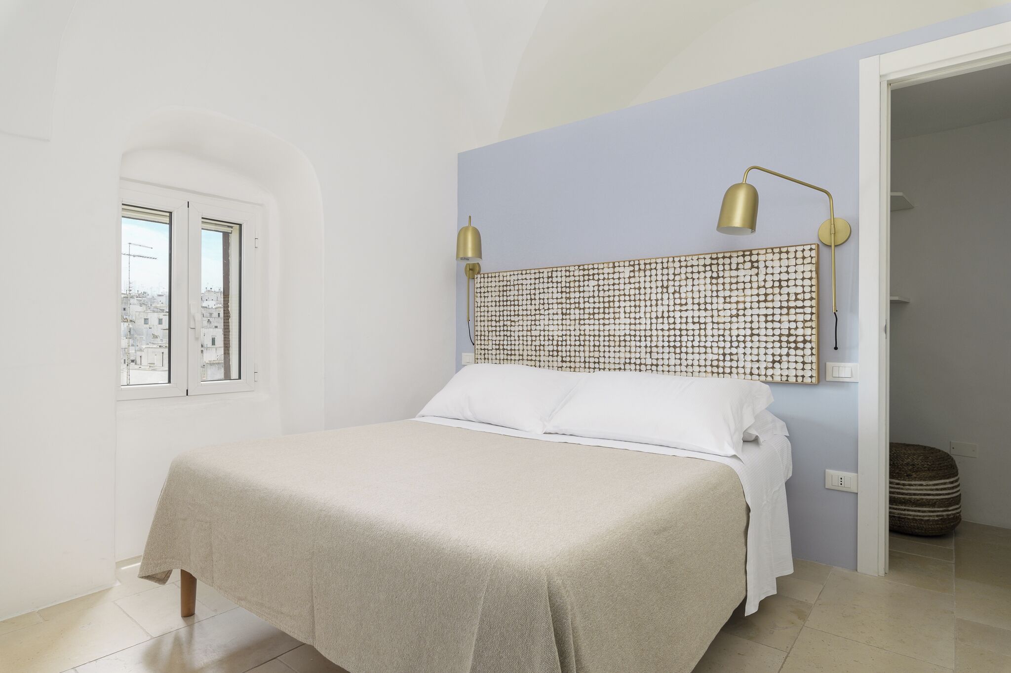 Un dormitorio con cama, dos lámparas de noche, cabecera estampada, ventana y vistas a edificios.