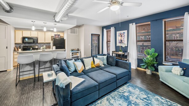 A modern living room with a blue sofa, a dining area with bar stools, a kitchen with a microwave, and a ceiling fan. - Near Wells Fargo, King Bed and Balcony and washer dryer, fitness room, Fireplace (Des Moines)